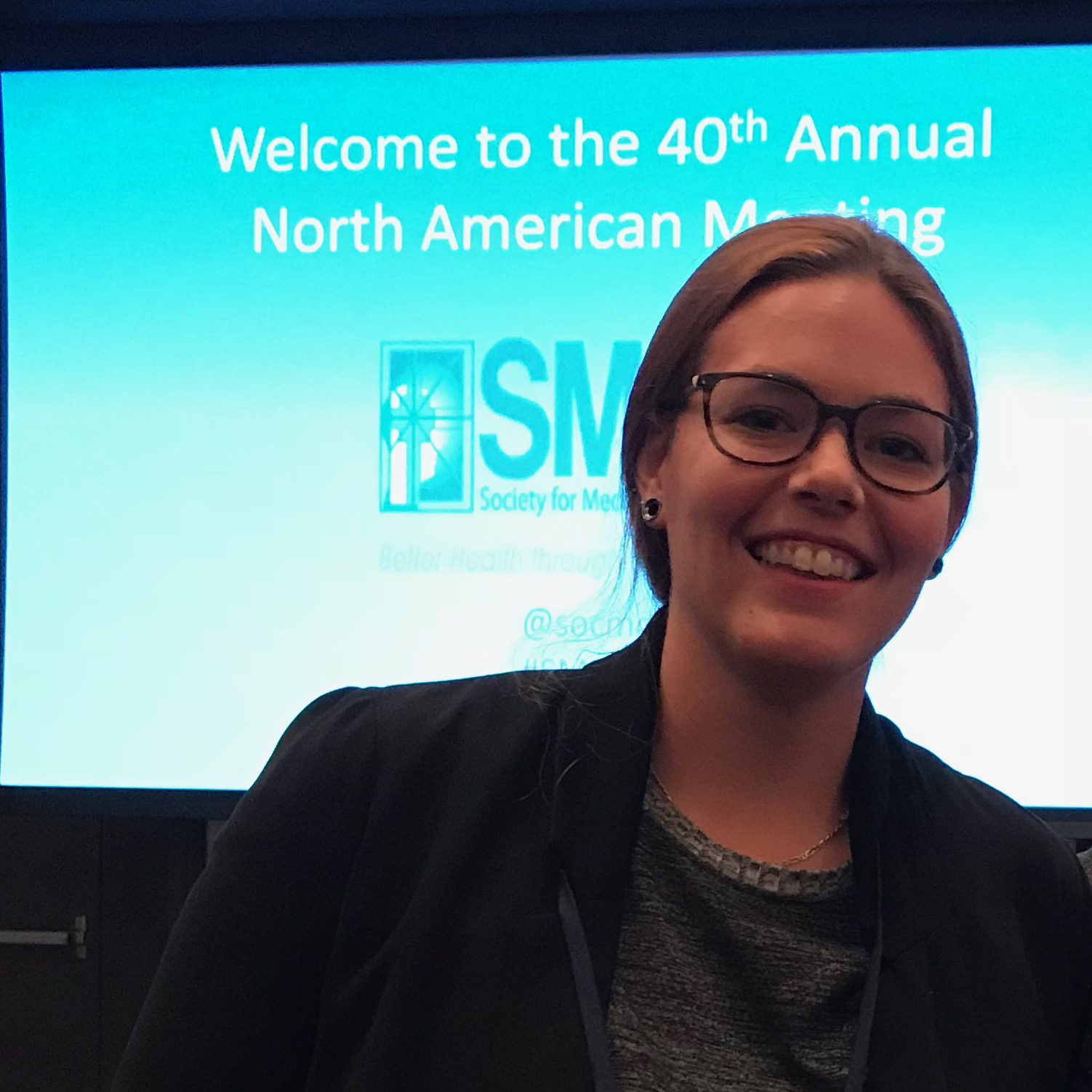 A smiling white woman in front of a conference screen